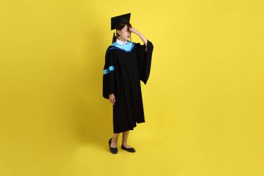 Sarı arka planda izole edilmiş stres jesti ile mavi mezuniyet elbisesi ve kepleri içinde bayan üniversite öğrencisi. Gen Z, yeni mezun, eğitim, ilk iş