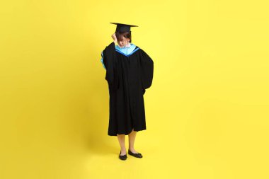 Sarı arka planda izole edilmiş stres jesti ile mavi mezuniyet elbisesi ve kepleri içinde bayan üniversite öğrencisi. Gen Z, yeni mezun, eğitim, ilk iş