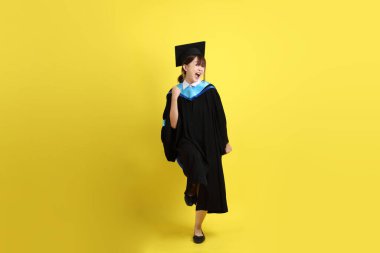 Mavi mezuniyet elbisesi ve şapkalı, heyecanlı, sarı arka planda izole bir şekilde kutlanan üniversite öğrencisi. Gen Z, yeni mezun, eğitim, ilk iş