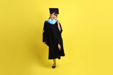 Mavi mezuniyet elbisesi ve şapkalı, heyecanlı, sarı arka planda izole bir şekilde kutlanan üniversite öğrencisi. Gen Z, yeni mezun, eğitim, ilk iş