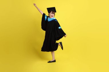 Mavi mezuniyet elbisesi ve şapkalı, heyecanlı, sarı arka planda izole bir şekilde kutlanan üniversite öğrencisi. Gen Z, yeni mezun, eğitim, ilk iş