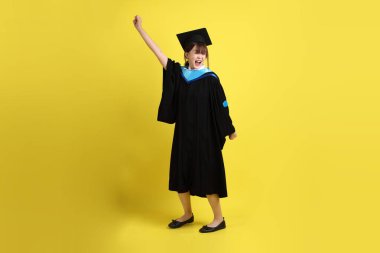 Mavi mezuniyet elbisesi ve şapkalı, heyecanlı, sarı arka planda izole bir şekilde kutlanan üniversite öğrencisi. Gen Z, yeni mezun, eğitim, ilk iş