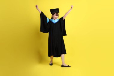 Mavi mezuniyet elbisesi ve şapkalı, heyecanlı, sarı arka planda izole bir şekilde kutlanan üniversite öğrencisi. Gen Z, yeni mezun, eğitim, ilk iş