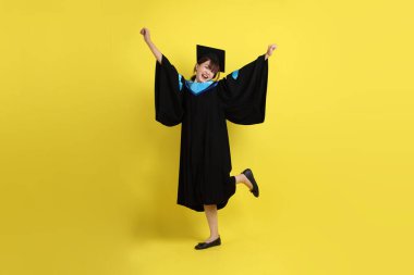 Mavi mezuniyet elbisesi ve şapkalı, heyecanlı, sarı arka planda izole bir şekilde kutlanan üniversite öğrencisi. Gen Z, yeni mezun, eğitim, ilk iş