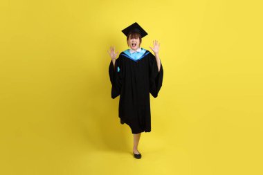 Mavi mezuniyet elbisesi ve şapkalı, heyecanlı, sarı arka planda izole bir şekilde kutlanan üniversite öğrencisi. Gen Z, yeni mezun, eğitim, ilk iş