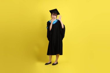 Mavi mezuniyet elbisesi ve şapkalı, sürpriz jestli ve sarı arka planda izole edilmiş etkileyici bir üniversite öğrencisi. Gen Z, yeni mezun, eğitim, ilk iş