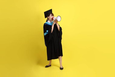 Mavi mezuniyet elbiseleri ve kepleri içinde elinde megafonla sarı arka plan izole bir şekilde bağıran bir üniversite öğrencisi. Gen Z, yeni mezun, eğitim, ilk iş