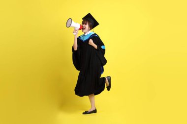 Mavi mezuniyet elbiseleri ve kepleri içinde elinde megafonla sarı arka plan izole bir şekilde bağıran bir üniversite öğrencisi. Gen Z, yeni mezun, eğitim, ilk iş