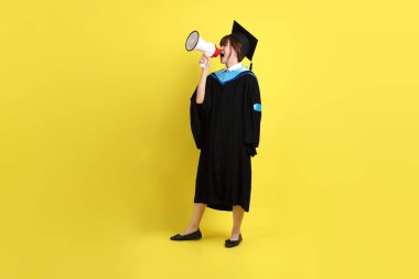 Mavi mezuniyet elbiseleri ve kepleri içinde elinde megafonla sarı arka plan izole bir şekilde bağıran bir üniversite öğrencisi. Gen Z, yeni mezun, eğitim, ilk iş