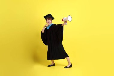 Mavi mezuniyet elbiseleri ve kepleri içinde elinde megafonla sarı arka plan izole bir şekilde bağıran bir üniversite öğrencisi. Gen Z, yeni mezun, eğitim, ilk iş