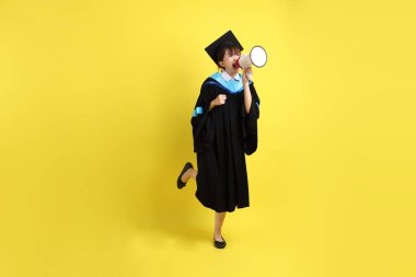 Mavi mezuniyet elbiseleri ve kepleri içinde elinde megafonla sarı arka plan izole bir şekilde bağıran bir üniversite öğrencisi. Gen Z, yeni mezun, eğitim, ilk iş