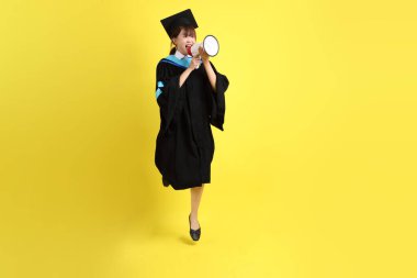 Mavi mezuniyet elbiseleri ve kepleri içinde elinde megafonla sarı arka plan izole bir şekilde bağıran bir üniversite öğrencisi. Gen Z, yeni mezun, eğitim, ilk iş