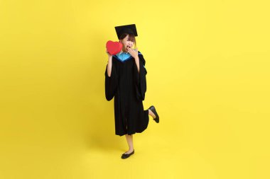 Mavi mezuniyet elbisesi ve kepleri takmış, kalp şeklinde jestleri olan, küçük kalbi olan, sarı arka planda izole bir aşk yaşayan bir üniversite öğrencisi. Gen Z, yeni mezun, eğitim, ilk iş