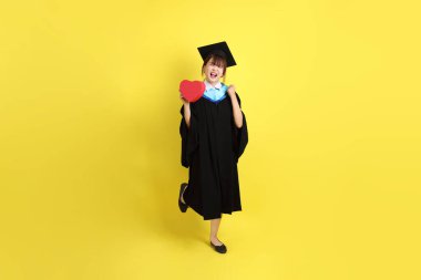 Mavi mezuniyet elbisesi ve kepleri takmış, kalp şeklinde jestleri olan, küçük kalbi olan, sarı arka planda izole bir aşk yaşayan bir üniversite öğrencisi. Gen Z, yeni mezun, eğitim, ilk iş