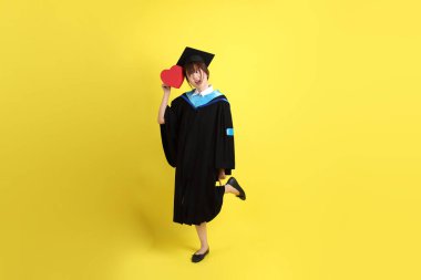 Mavi mezuniyet elbisesi ve kepleri takmış, kalp şeklinde jestleri olan, küçük kalbi olan, sarı arka planda izole bir aşk yaşayan bir üniversite öğrencisi. Gen Z, yeni mezun, eğitim, ilk iş