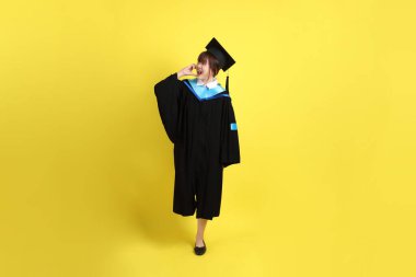 Mavi mezuniyet elbisesi ve kepleri takmış, kalp şeklinde jestleri olan, küçük kalbi olan, sarı arka planda izole bir aşk yaşayan bir üniversite öğrencisi. Gen Z, yeni mezun, eğitim, ilk iş