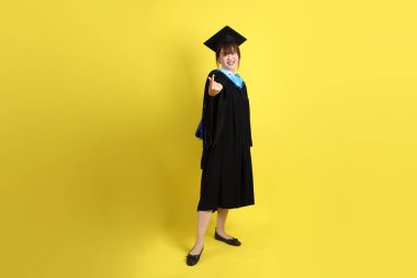 Mavi mezuniyet elbisesi ve kepleri takmış, kalp şeklinde jestleri olan, küçük kalbi olan, sarı arka planda izole bir aşk yaşayan bir üniversite öğrencisi. Gen Z, yeni mezun, eğitim, ilk iş