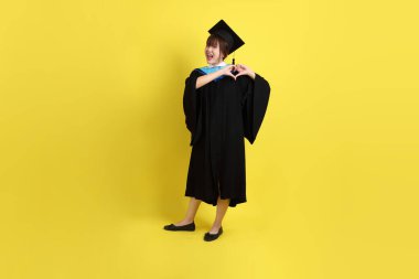 Mavi mezuniyet elbisesi ve kepleri takmış, kalp şeklinde jestleri olan, küçük kalbi olan, sarı arka planda izole bir aşk yaşayan bir üniversite öğrencisi. Gen Z, yeni mezun, eğitim, ilk iş