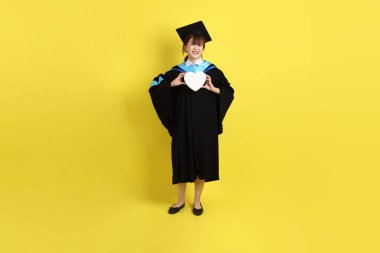 Mavi mezuniyet elbisesi ve kepleri takmış, kalp şeklinde jestleri olan, küçük kalbi olan, sarı arka planda izole bir aşk yaşayan bir üniversite öğrencisi. Gen Z, yeni mezun, eğitim, ilk iş