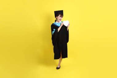 Mavi mezuniyet elbisesi ve kepleri takmış, kalp şeklinde jestleri olan, küçük kalbi olan, sarı arka planda izole bir aşk yaşayan bir üniversite öğrencisi. Gen Z, yeni mezun, eğitim, ilk iş