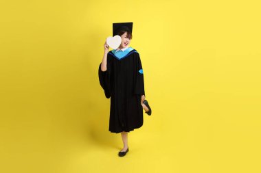 Mavi mezuniyet elbisesi ve kepleri takmış, kalp şeklinde jestleri olan, küçük kalbi olan, sarı arka planda izole bir aşk yaşayan bir üniversite öğrencisi. Gen Z, yeni mezun, eğitim, ilk iş