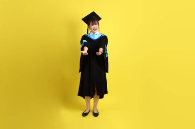 Mavi mezuniyet elbisesi ve kepleri içinde, sarı arka planda izole edilmiş, verme ve alma hareketi yapan bir üniversite öğrencisi. Gen Z, yeni mezun, eğitim, ilk iş