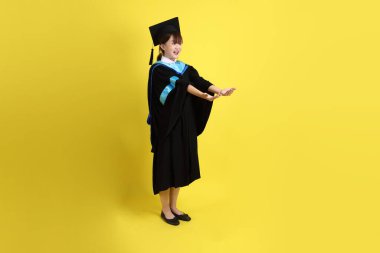 Mavi mezuniyet elbisesi ve kepleri içinde, sarı arka planda izole edilmiş, verme ve alma hareketi yapan bir üniversite öğrencisi. Gen Z, yeni mezun, eğitim, ilk iş