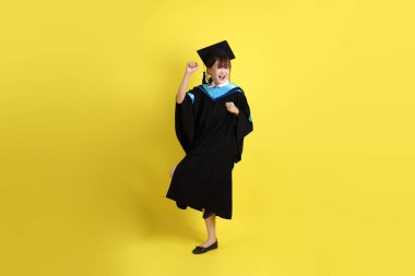 Mavi mezuniyet elbisesi ve şapkalı, heyecanlı, sarı arka planda izole bir şekilde kutlanan üniversite öğrencisi. Gen Z, yeni mezun, eğitim, ilk iş