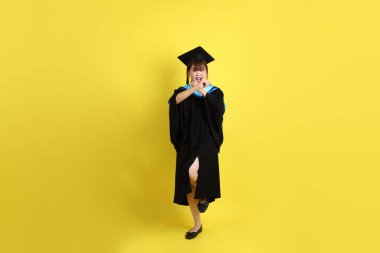 Mavi mezuniyet elbisesi ve şapkalı, sarı arka planda izole edilmiş dövüş jesti olan bir üniversite öğrencisi. Gen Z, yeni mezun, eğitim, ilk iş