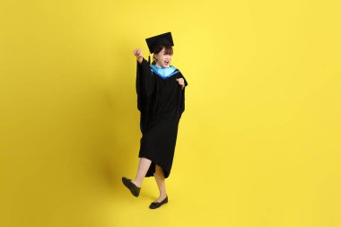 Mavi mezuniyet elbisesi ve şapkalı, sarı arka planda izole edilmiş dövüş jesti olan bir üniversite öğrencisi. Gen Z, yeni mezun, eğitim, ilk iş