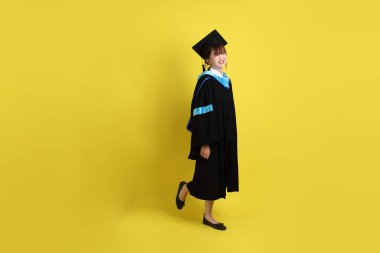 Mavi mezuniyet elbisesi ve kepli, sarı arka planda yalıtılmış yürüme hareketi yapan bir üniversite öğrencisi. Gen Z, yeni mezun, eğitim, ilk iş