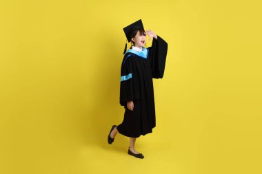 Mavi mezuniyet elbisesi ve şapkalı, sarı arka planda izole edilmiş mesafeye bakma hareketi yapan bir üniversite öğrencisi. Gen Z, yeni mezun, eğitim, ilk iş