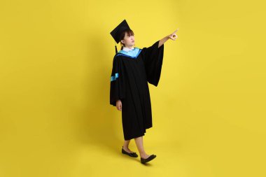 Mavi mezuniyet elbisesi ve kepleri içinde, sarı arka planda izole edilmiş bir şekilde işaret eden bayan üniversite öğrencisi. Gen Z, yeni mezun, eğitim, ilk iş