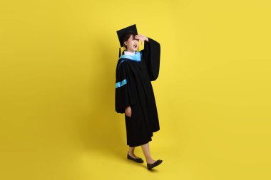 Mavi mezuniyet elbisesi ve şapkalı, sarı arka planda izole edilmiş mesafeye bakma hareketi yapan bir üniversite öğrencisi. Gen Z, yeni mezun, eğitim, ilk iş