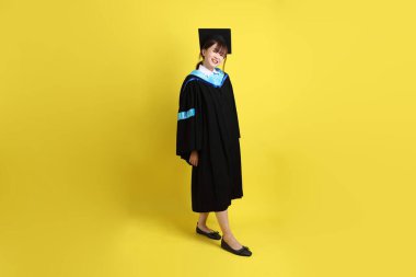 Mavi mezuniyet elbisesi ve kepli, sarı arka planda yalıtılmış yürüme hareketi yapan bir üniversite öğrencisi. Gen Z, yeni mezun, eğitim, ilk iş
