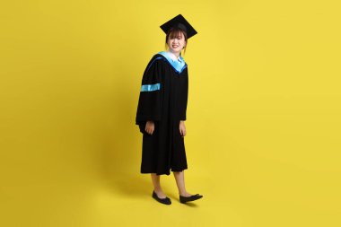 Mavi mezuniyet elbisesi ve kepli, sarı arka planda yalıtılmış yürüme hareketi yapan bir üniversite öğrencisi. Gen Z, yeni mezun, eğitim, ilk iş