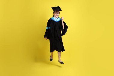 Sarı arka planda izole edilmiş mavi mezuniyet elbiseleri ve kepleri içinde atlama jesti olan bir üniversite öğrencisi. Gen Z, yeni mezun, eğitim, ilk iş