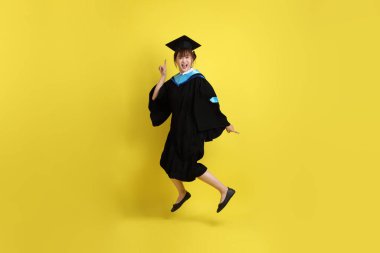 Sarı arka planda izole edilmiş mavi mezuniyet elbiseleri ve kepleri içinde atlama jesti olan bir üniversite öğrencisi. Gen Z, yeni mezun, eğitim, ilk iş