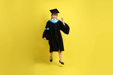 Sarı arka planda izole edilmiş mavi mezuniyet elbiseleri ve kepleri içinde atlama jesti olan bir üniversite öğrencisi. Gen Z, yeni mezun, eğitim, ilk iş