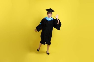 Mavi mezuniyet elbisesi ve şapkalı, sürpriz jestli ve sarı arka planda izole edilmiş etkileyici bir üniversite öğrencisi. Gen Z, yeni mezun, eğitim, ilk iş