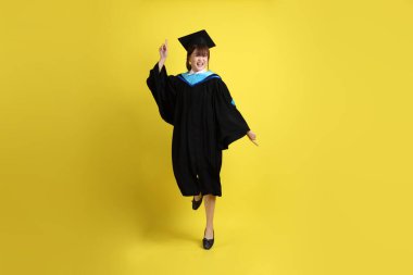 Sarı arka planda izole edilmiş mavi mezuniyet elbiseleri ve kepleri içinde atlama jesti olan bir üniversite öğrencisi. Gen Z, yeni mezun, eğitim, ilk iş