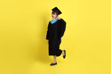 Mavi mezuniyet elbisesi ve kepleri içinde sarı arka planda izole Çağrı jesti taşıyan bir üniversite öğrencisi. Gen Z, yeni mezun, eğitim, ilk iş