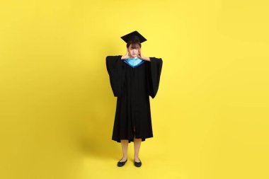 Sarı arka planda, kulağı kapalı, mavi mezuniyet elbisesi ve şapkası takmış bir üniversite öğrencisi. Gen Z, yeni mezun, eğitim, ilk iş