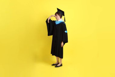 Mavi mezuniyet elbisesi ve kepli, koklama jestli, sarı arka planda kapalı burunlu bir üniversite öğrencisi. Gen Z, yeni mezun, eğitim, ilk iş