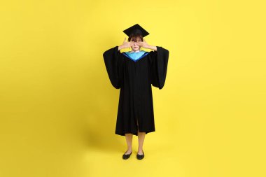 Mavi mezuniyet elbisesi ve şapkalı, sarı arka planda izole edilmiş örtülü gözleri olan bir üniversite öğrencisi. Gen Z, yeni mezun, eğitim, ilk iş