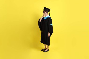 Mavi mezuniyet elbisesi ve kepli, koklama jestli, sarı arka planda kapalı burunlu bir üniversite öğrencisi. Gen Z, yeni mezun, eğitim, ilk iş