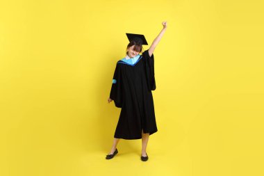 Mavi mezuniyet elbisesi ve şapkalı, heyecanlı, sarı arka planda izole bir şekilde kutlanan üniversite öğrencisi. Gen Z, yeni mezun, eğitim, ilk iş