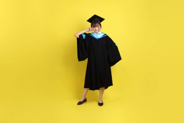 Mavi mezuniyet elbisesi ve şapkalı bayan üniversite öğrencisi. Sarı arka planda uykulu ya da esnemiş jestleri var. Gen Z, yeni mezun, eğitim, ilk iş