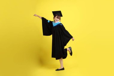 Mavi mezuniyet elbisesi ve şapkalı, heyecanlı, sarı arka planda izole bir şekilde kutlanan üniversite öğrencisi. Gen Z, yeni mezun, eğitim, ilk iş