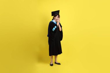 Sarı arka planda hastalıklı bir jestle mavi mezuniyet elbisesi ve kepleri olan bir üniversite öğrencisi. Gen Z, yeni mezun, eğitim, ilk iş